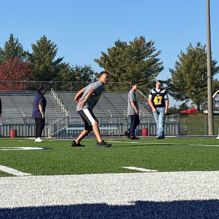 Four adults on a football field, three wearing jerseys, one wearing a blue shirt, with bleachers and trees in the background.