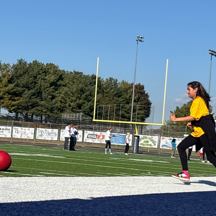 A football field with a ball on the ground, a person in a yellow shirt, and several others standing around.