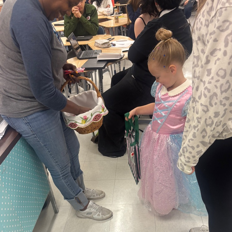 Woman holding basket hands it to young girl in pink dress, who holds black bag. People seated in background.