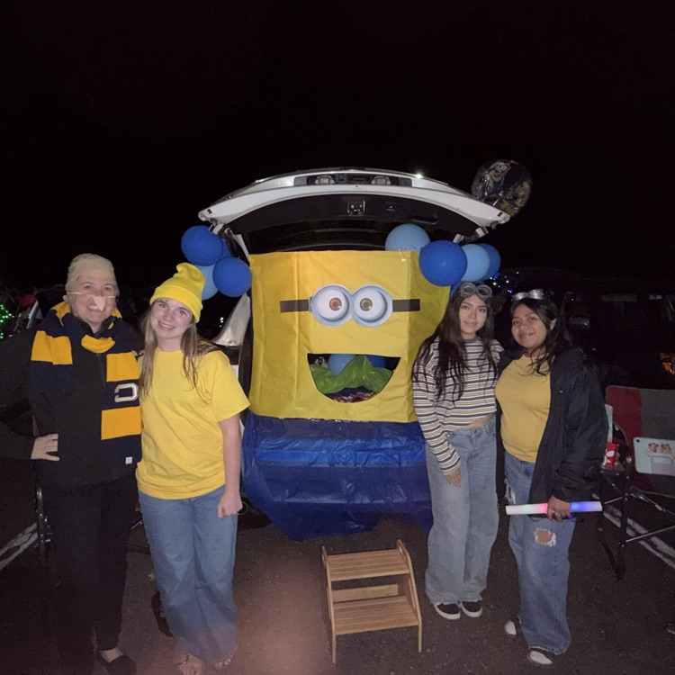 Four people wearing yellow shirts stand in front of a Minions float at night.