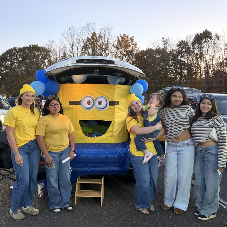 Group of people standing in front of a car with a minion inflatable, all wearing matching yellow shirts.