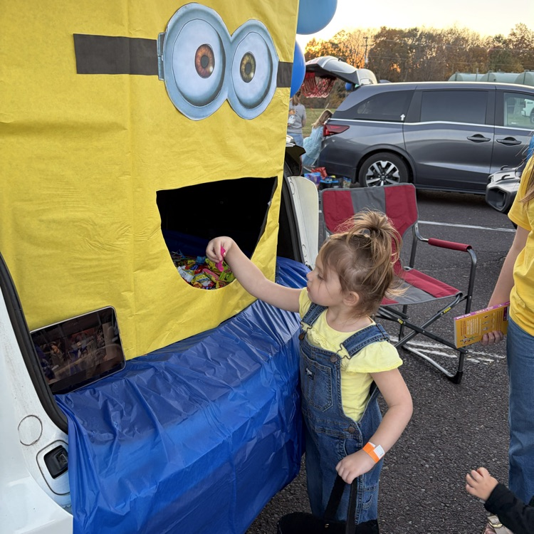 A child wearing a yellow shirt and overalls collects candy from a large yellow costumed mascot in a parking lot.