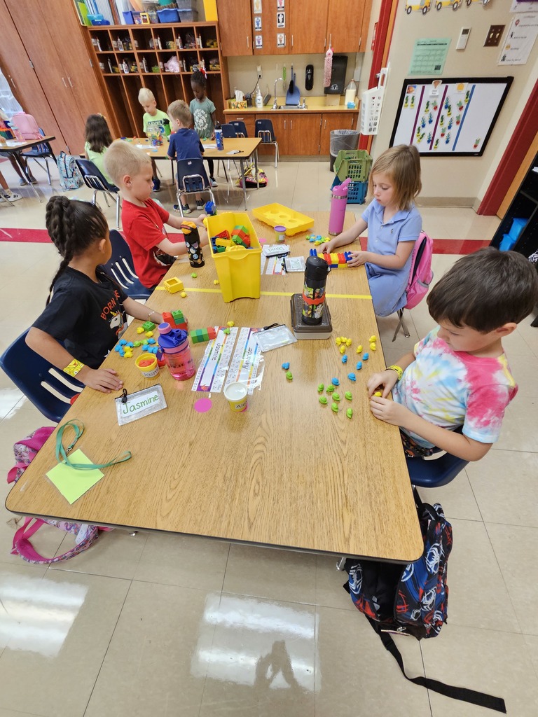 a group of four kindergartners stands around a rectangular table and explore various stem tools such as play-doh, and Duplo blocks
