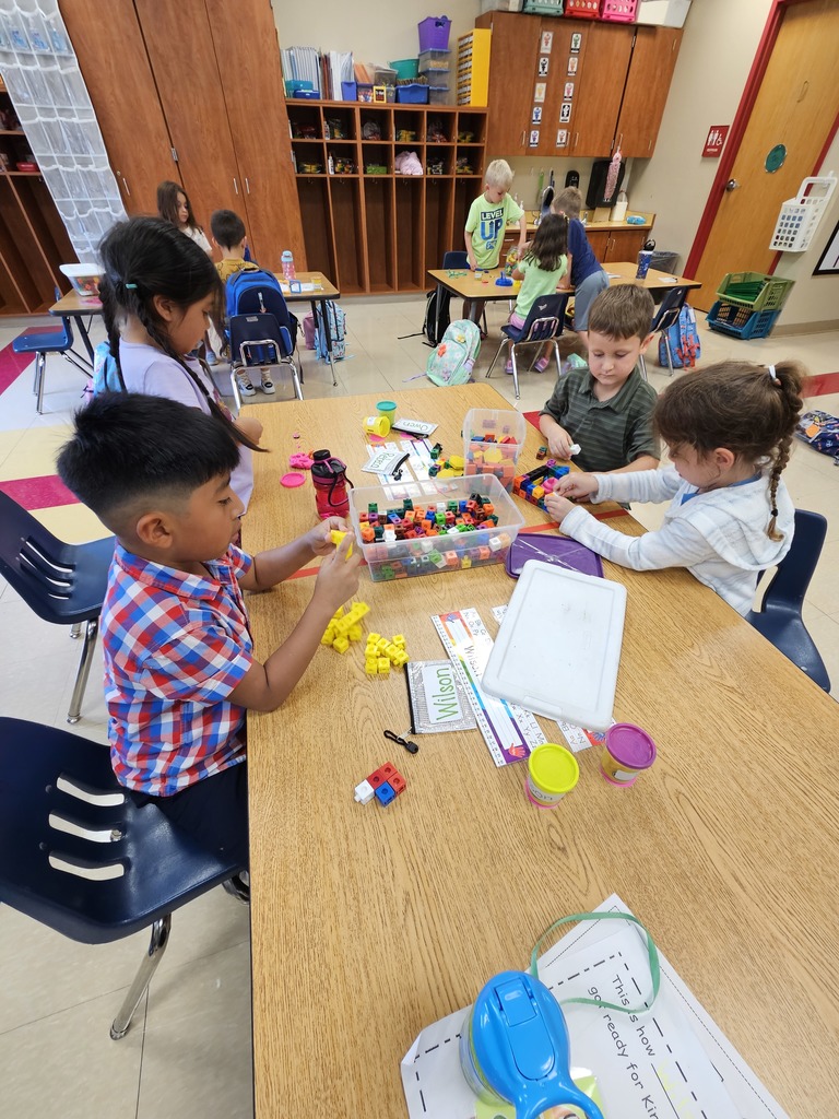 a group of four kindergartners stands around a rectangular table and explore various stem tools such as play-doh, and Duplo blocks
