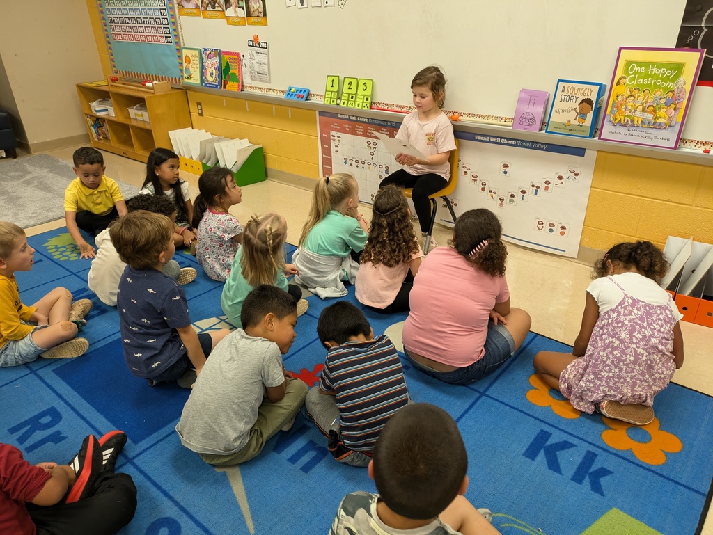 One kindergarten student sits in a chair in the front of the classroom and reads her self created book to the class.