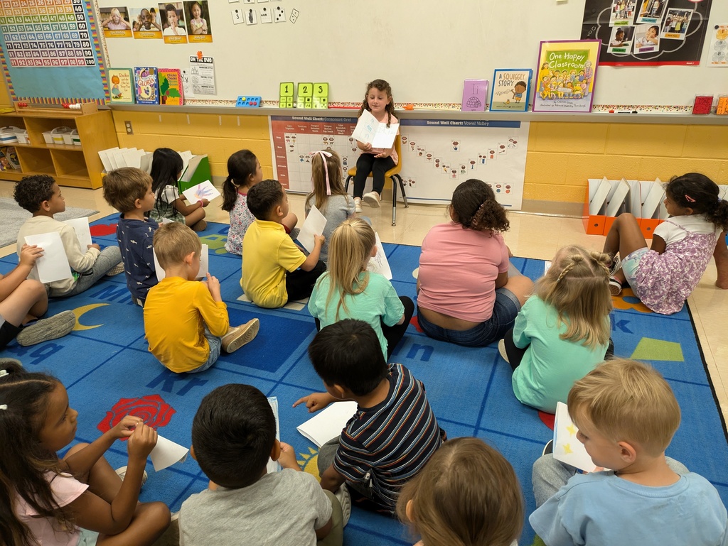 One kindergarten student sits in a chair in the front of the classroom and reads her self created book to the class.
