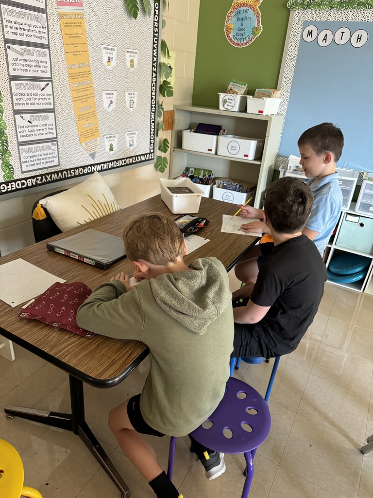 three third grade boys sit on colorful stools at a table in a third grade classroom and work on a project together. 