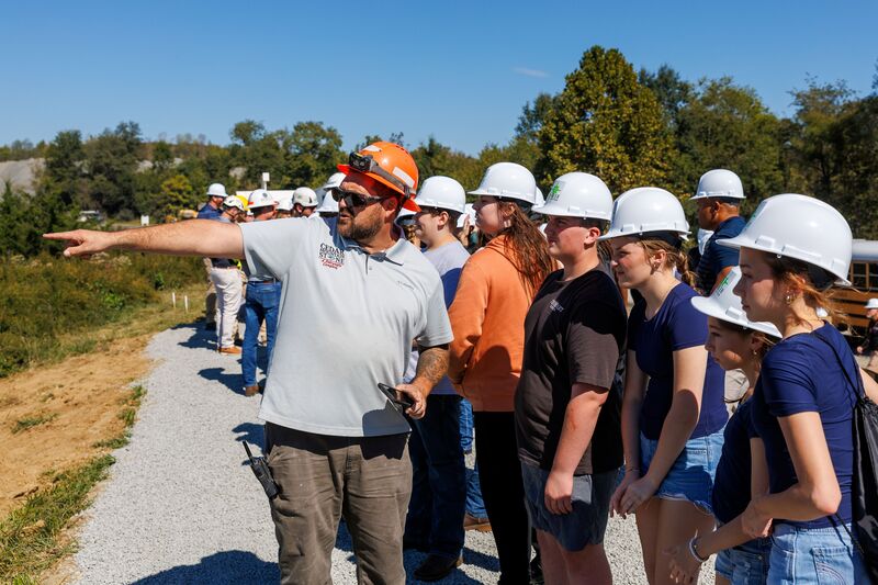 A construction professional wearing an orange hard hat points toward a worksite while speaking to a group of students wearing white hard hats during a career exploration visit.