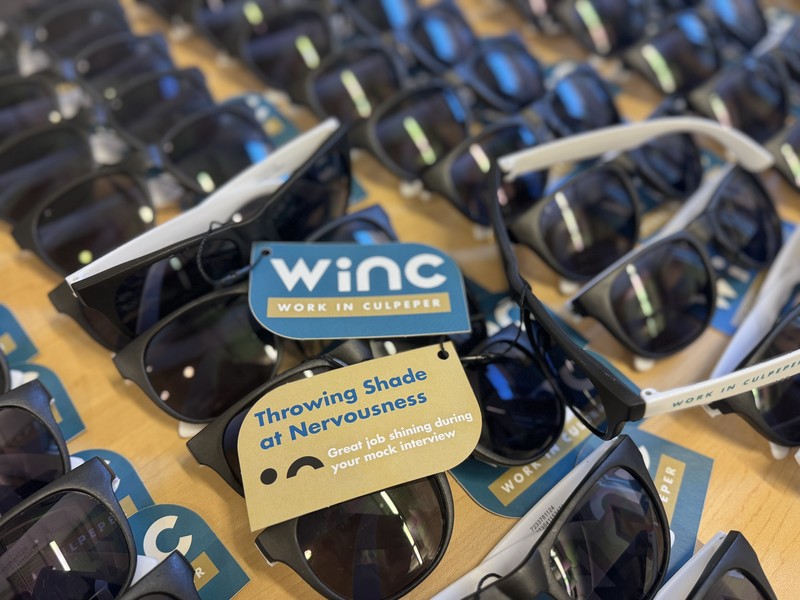 A close-up of rows of black sunglasses laid out on a table, each tagged with a blue and tan card that reads “Throwing Shade at Nervousness – Great job shining during your mock interview.” The tags also feature the Work in Culpeper (WiNC) logo.