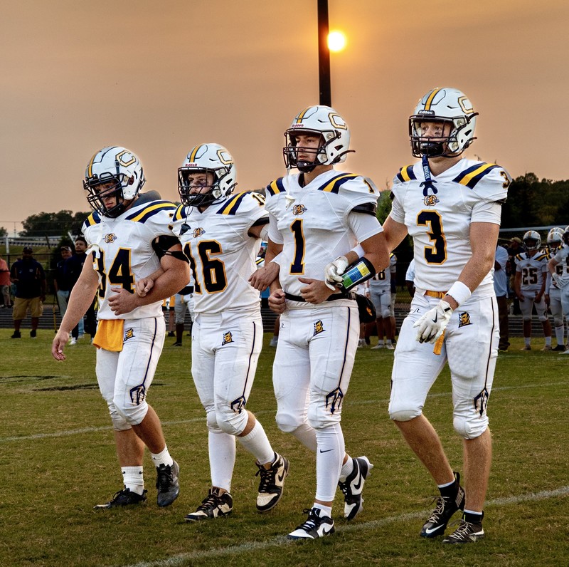 Four Culpeper County High School football players in white uniforms with blue and gold accents walk arm in arm across the field at sunset. They wear helmets and game gear, with the sun glowing in the background and teammates visible behind them.