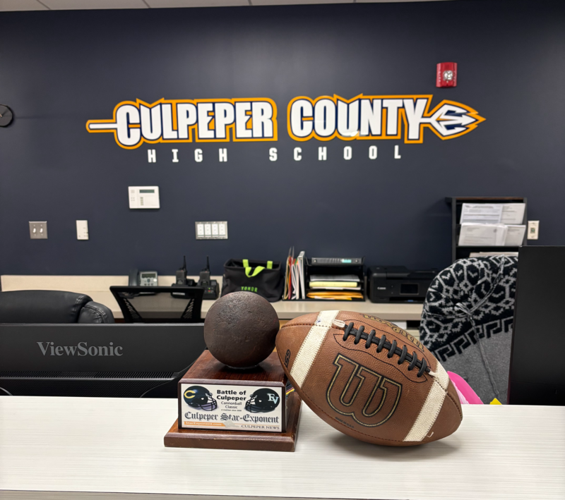 A football and the Cannonball Classic trophy sit on a desk inside Culpeper County High School. The wall behind the desk displays a large Culpeper County High School logo in blue and gold.