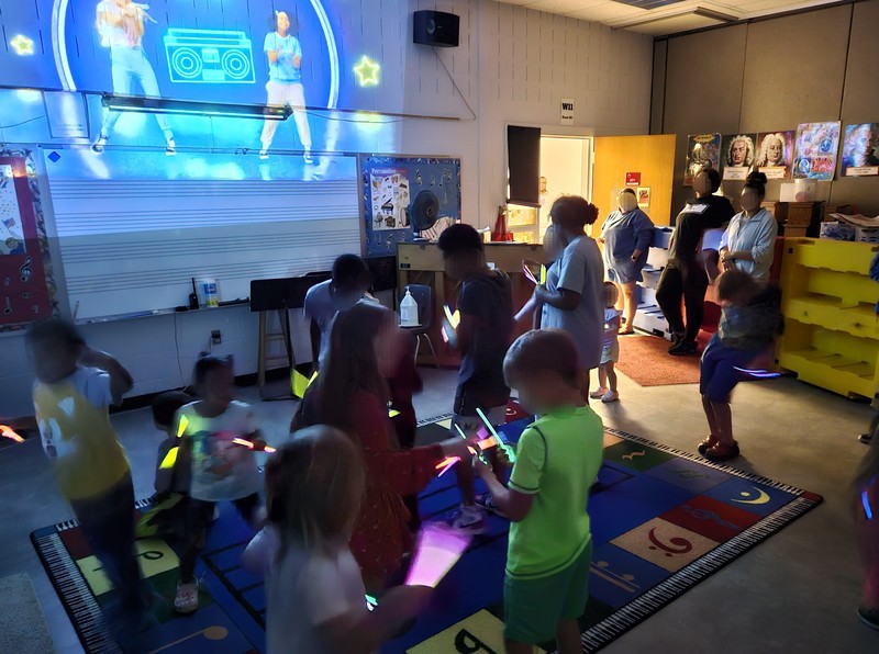 Students and families dance with glow sticks in a darkened music classroom at AG Richardson Elementary during Back to School Night. A colorful video with dancers and a boombox is projected on the wall, creating a lively dance party atmosphere.