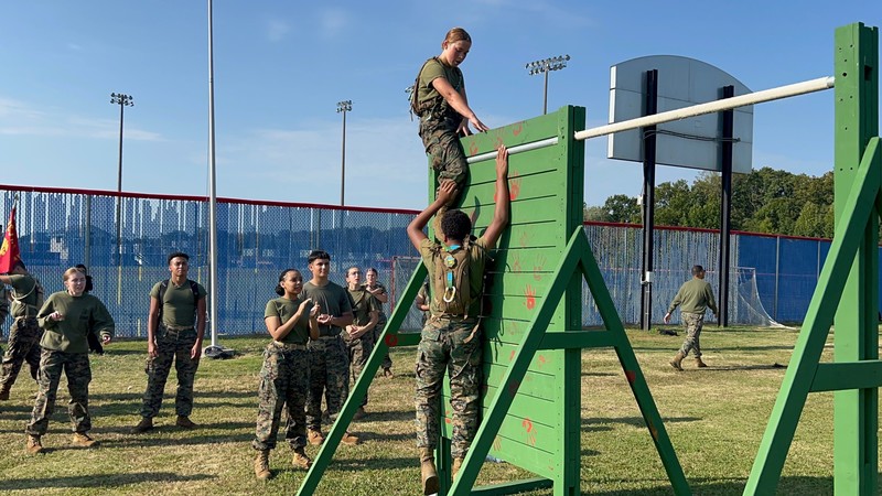 MCJROTC cadet climbs over a wooden wall in an obstacle course while teammate assists and others look on