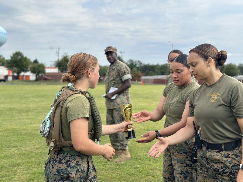 mcjrotc cadet hands trophy to 2 other cadets from another school on a sunny day while adult looks on