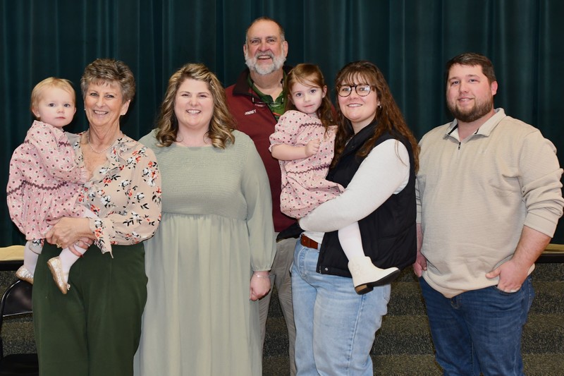 Maci Lane stands with her parents, husband, sister and two young daughters in front of the school stage curtain