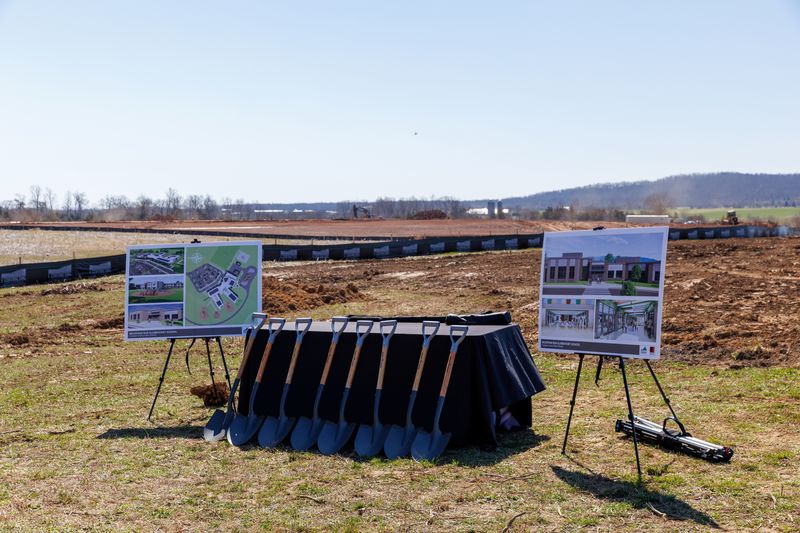 table with 8 shovels leaned against it with renderings of a school building on either side with construction site in the background