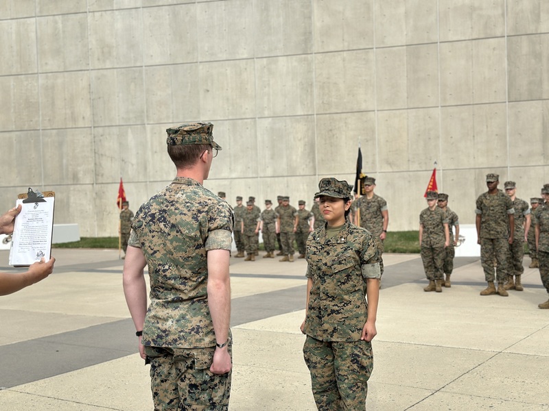 Two JROTC cadets in camouflage uniforms stand facing each other at attention during a formal inspection or ceremony, while additional cadets stand in formation behind them; a person at the left holds a clipboard with paperwork.