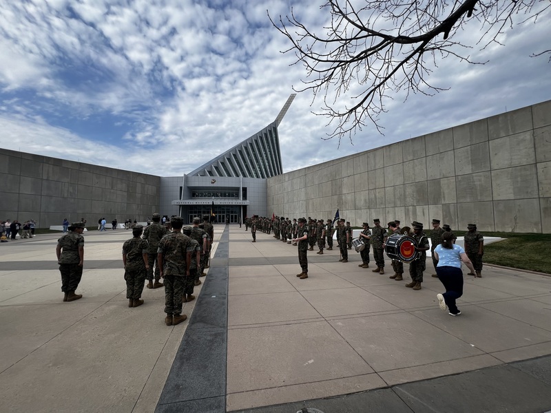 A large formation of JROTC cadets stands in organized rows outside the National Museum of the Marine Corps, with a drumline positioned to the right and the museum’s distinctive angular structure rising in the background under a partly cloudy sky.