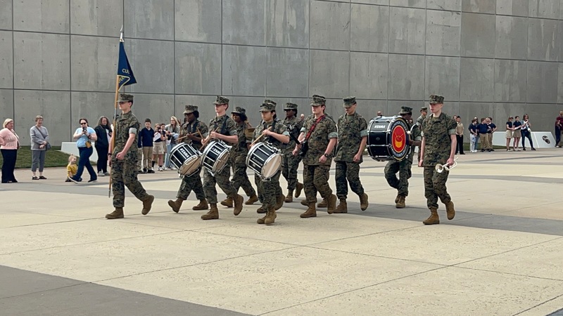 A group of JROTC cadets in camouflage uniforms marches in formation across a plaza, playing drums and carrying a flag, while a crowd of onlookers stands nearby against a large concrete wall.