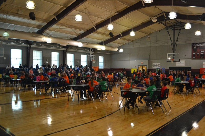 school gym filled with tables of 4 students and a teacher for math 24 competition with spectators in the bleachers behind them