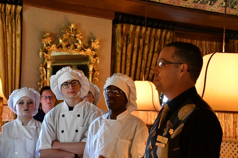 Culinary Arts students in white chef uniforms listen attentively to a staff member speaking in an elegant dining room at The Inn at Little Washington, with warm lighting, decorative mirrors, and draped curtains in the background.