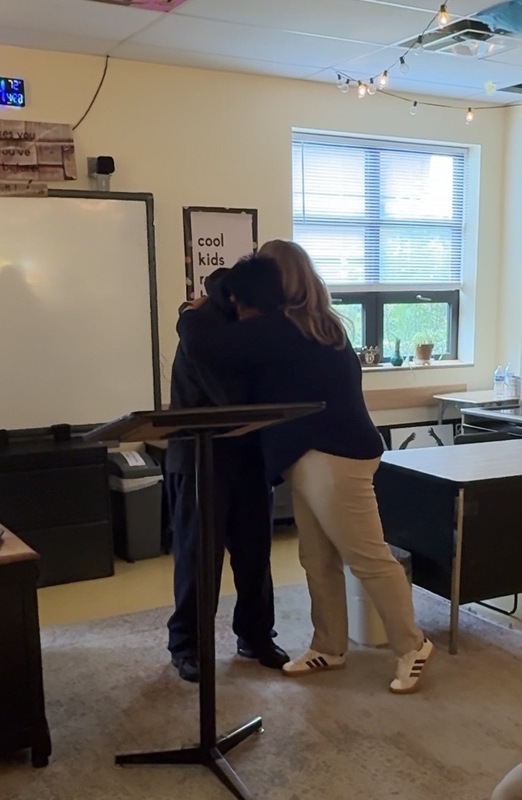 student in classroom hugging teacher