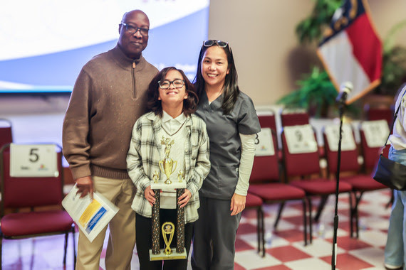 CCS' 2026 Spelling Bee Champ Angelika Johnson takes a quick photo with her parents while proudly holding her coveted trophy.