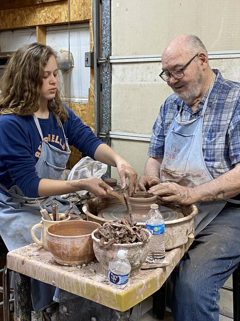 An art student is working with a teacher to throw clay on the wheel.