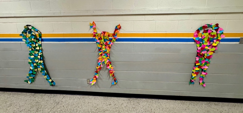Three black silhouette figures on a school hallway wall, each filled with folded paper shapes. The left figure is covered in green, blue, and yellow hearts; the middle is decorated with multicolored triangles and has arms raised; the right is filled with layered hearts in pink, red, teal, and yellow.