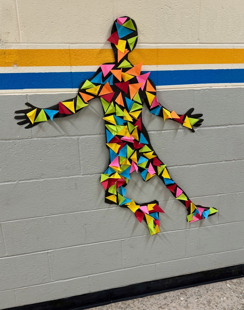 Silhouette of a person jumping, made from black paper and decorated with brightly colored, folded triangle shapes in pink, yellow, blue, green, and orange. The figure is mounted on a gray cinderblock wall with horizontal yellow, white, and blue stripes.