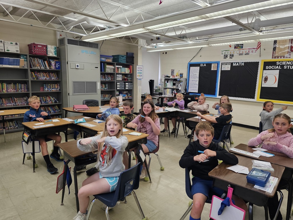 Students are actively engaging with the prefix "un-" and the Greek roots "graph" and "gram" through interactive hand motions, reinforcing their learning.