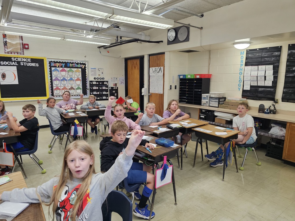 Students are actively engaging with the prefix "un-" and the Greek roots "graph" and "gram" through interactive hand motions, reinforcing their learning.