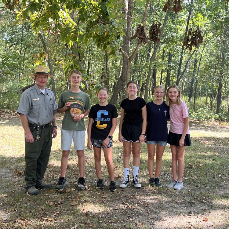 The team is standing in the woods with one of the Rangers. They are holding their acorn trophy.