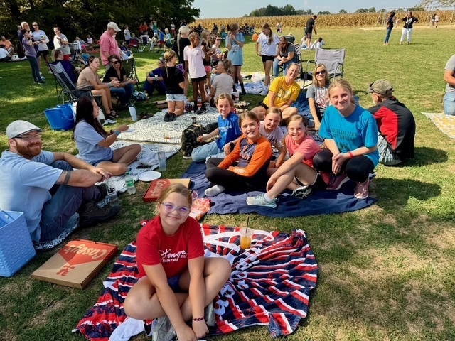Families are sitting together on the lawn in groups. They are talking and eating their lunch.