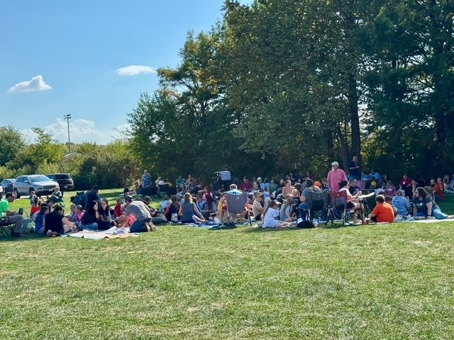 Families are sitting together on the lawn in groups. They are talking and eating their lunch.