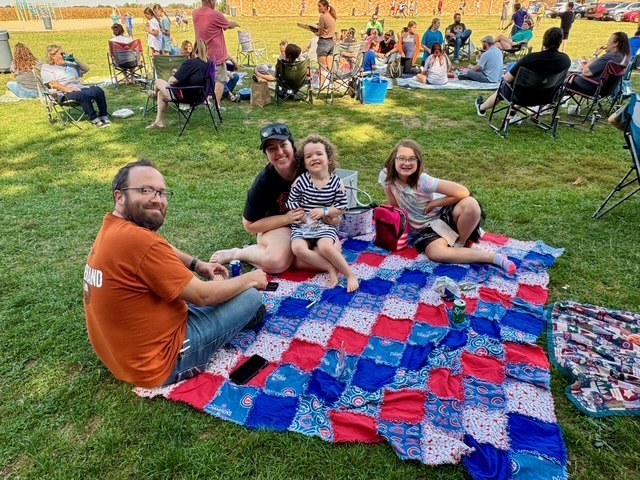 Families are sitting together on the lawn in groups. They are talking and eating their lunch.