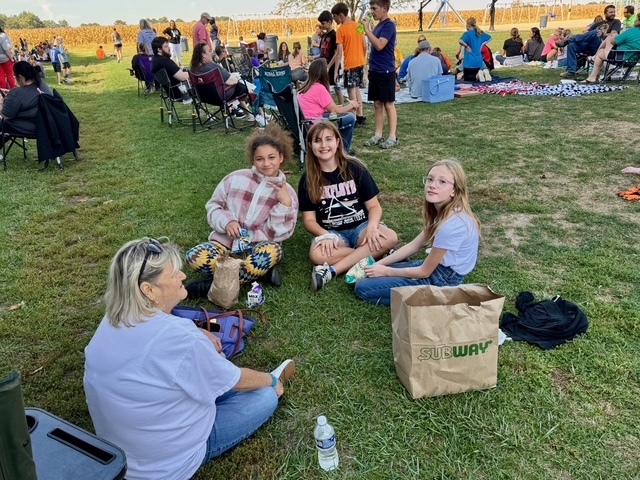 Families are sitting together on the lawn in groups. They are talking and eating their lunch.