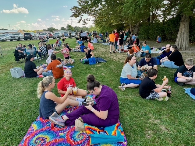Families are sitting together on the lawn in groups. They are talking and eating their lunch.