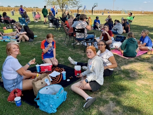 Families are sitting together on the lawn in groups. They are talking and eating their lunch.