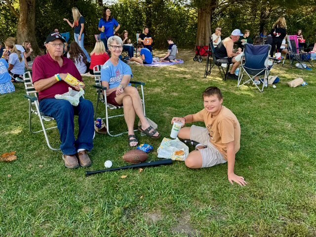 Families are sitting together on the lawn in groups. They are talking and eating their lunch.