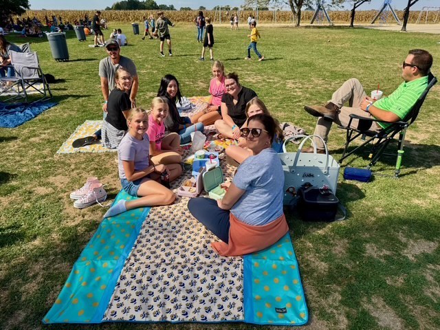 Families are sitting together on the lawn in groups. They are talking and eating their lunch.