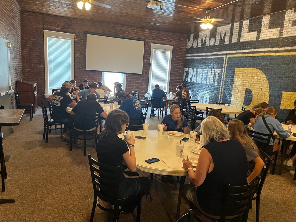 Students are sitting around round tables talking and eating their pizza.