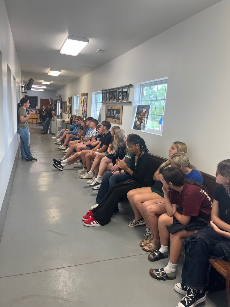 Students are sitting along the same wall on a very long bench. They are listening to a lady speak about how they make cheese.