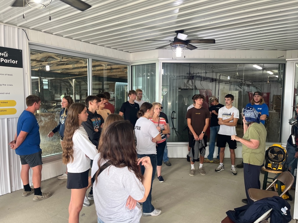 Students are gathered around a metal room with large windows. A lady is speaking to them about the process they use to make cheese.