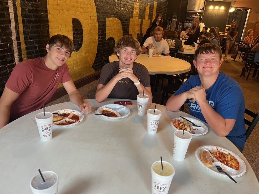 Students are sitting around a round table. They are eating thier pasta.