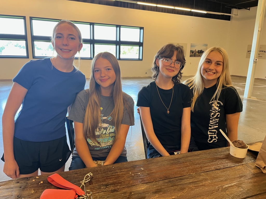 Four students are standing behind a table. They are in a large white room.