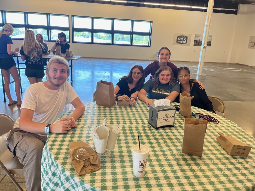 Students are sitting around a round table with a checkerboard tablecloth. They are posing together with thier empty bags on the table.