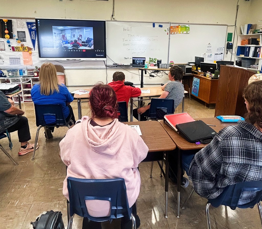 The class is talking with someone in another time zone via Google Meet. They are facing the Smartboard and asking them questions.