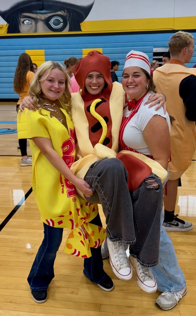 Three students are together. Two are holding the third student. The first is a sugar daddy candy, the second is a hot dog, and the third is a concession stand worker.