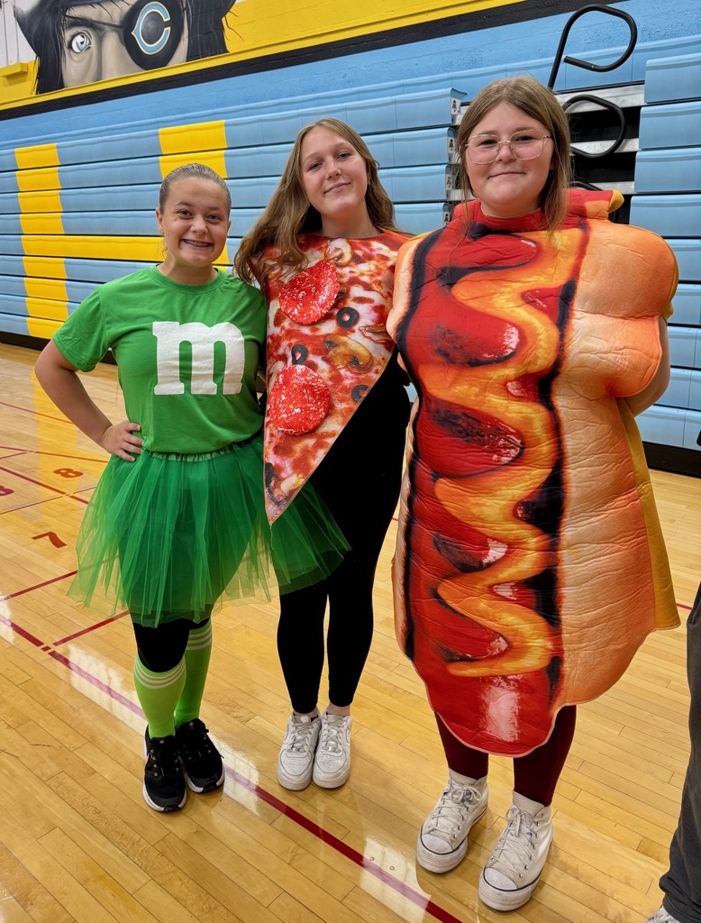 Three students are standing side by side. The first is a green m&m. She is wearing a green skirt, green socks and green shirt with an m. The second is wearing a pizza costume. The third is wearing a hotdog costume.